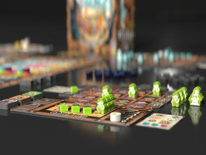 A close-up of a board game setup on a dark reflective surface, featuring a brown game board with green player tokens, cards, and stacks of small pieces in the foreground, with a blurred colorful game box in the background.
