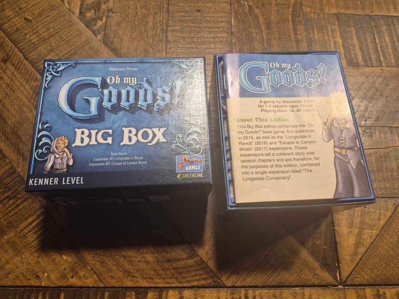 The 'Oh My Goods! Big Box' board game and its instruction booklet are displayed on a dark wood parquet floor. The packaging features a dark blue and silver design with white gothic lettering, and a small 'Kings of the Coast' logo is visible on the top right.