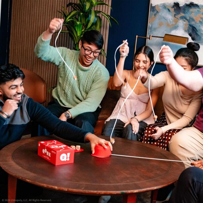 Group of people playing a game around a wooden table with a red box labeled 'Q!' in a casual setting.