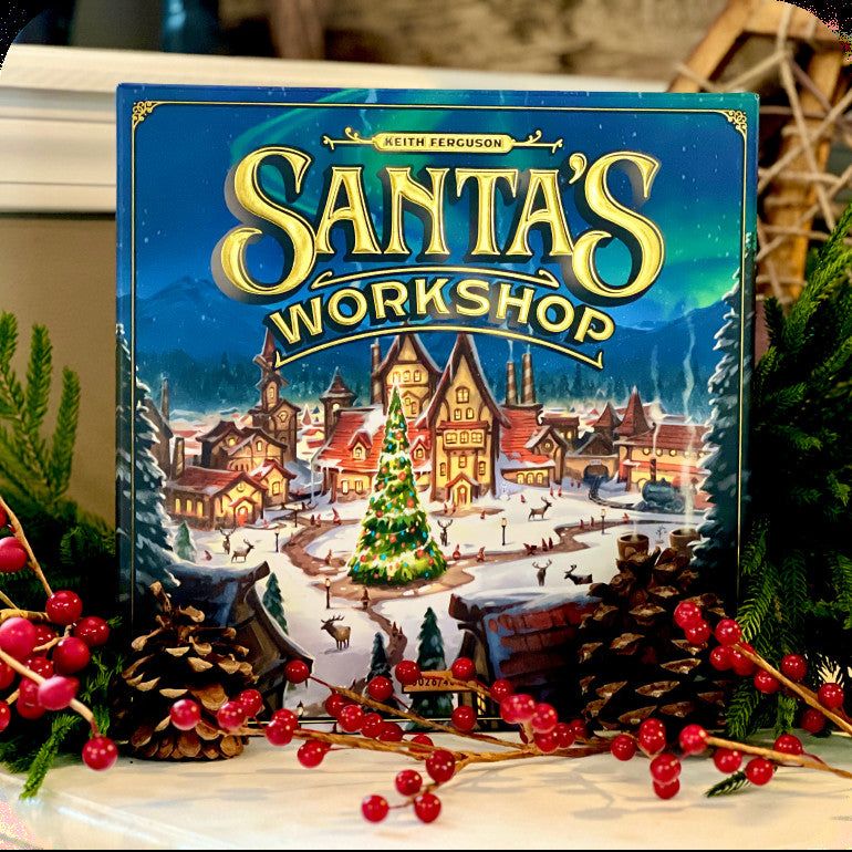 The board game box for 'Santa's Workshop' by Keith Ferguson is displayed on a tabletop decorated with pine branches, pinecones, and red berries. The box features a festive winter village scene with a glowing Christmas tree under a teal night sky, with the title in large gold letters.