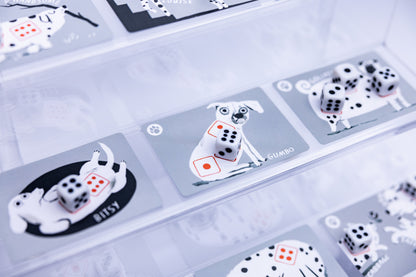 A close-up of a clear display case containing several gray cards with illustrated white dogs and black-and-white dice, some with small red accents. The cards are arranged on a light surface, and the photo has a soft, slightly hazy aesthetic.