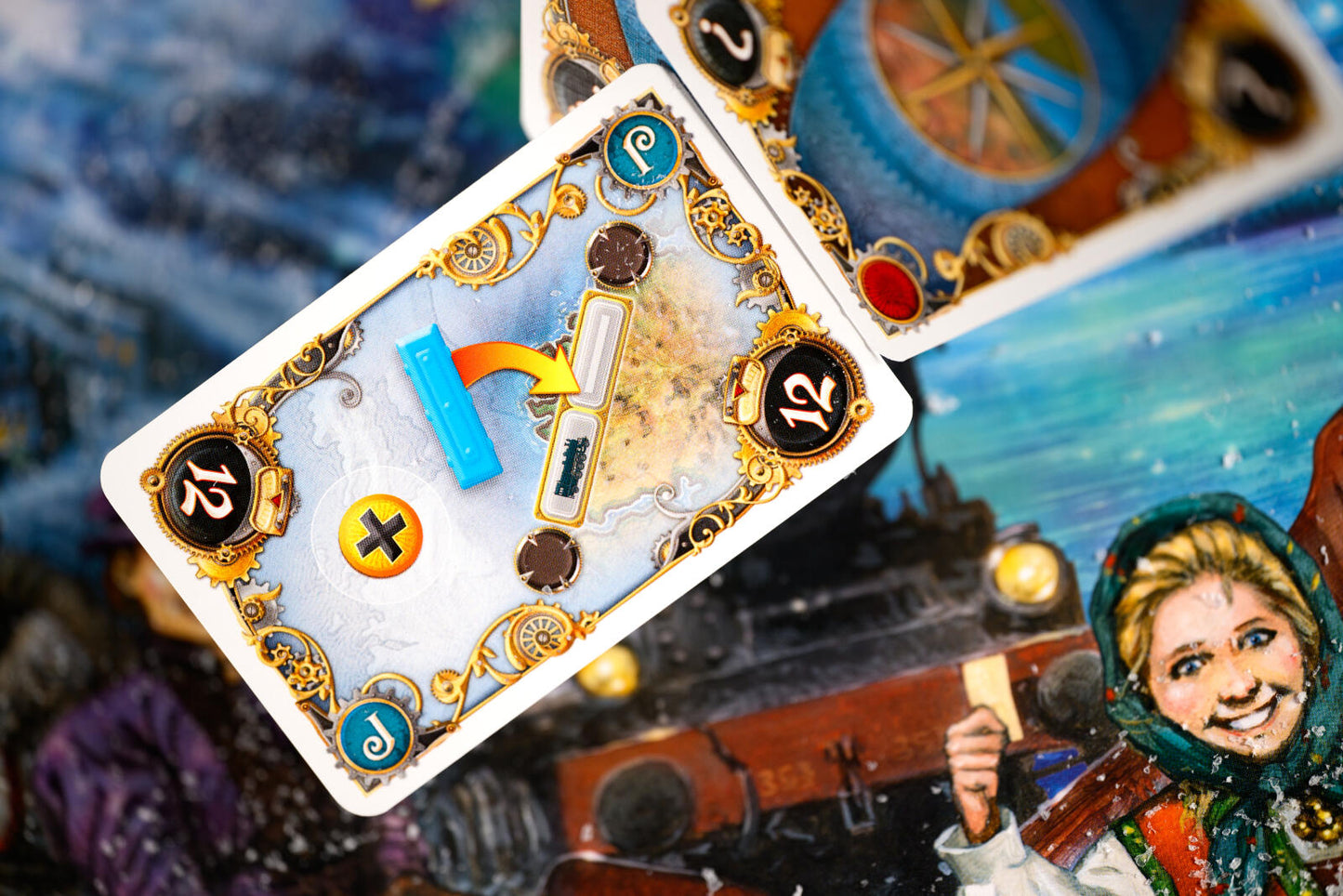Ticket to ride northern lights with a close-up of a decorative playing card with ornate gold detailing, blue and yellow symbols, and the number 12, layered over another illustrated card featuring a woman in a teal headscarf against a snowy blue-toned background.