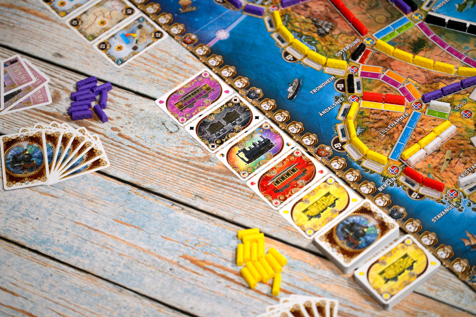Ticket to ride northern lights close-up of a colorful board game spread across a rustic light blue wooden table. Visible elements include a blue ocean-themed game board, multiple player cards, small purple and yellow tokens, and several fanned cards.