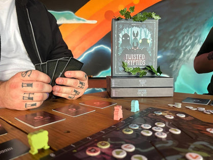 Board game setup with 'Twisted Cryptids' box and player hands holding cards, against a colorful abstract background.