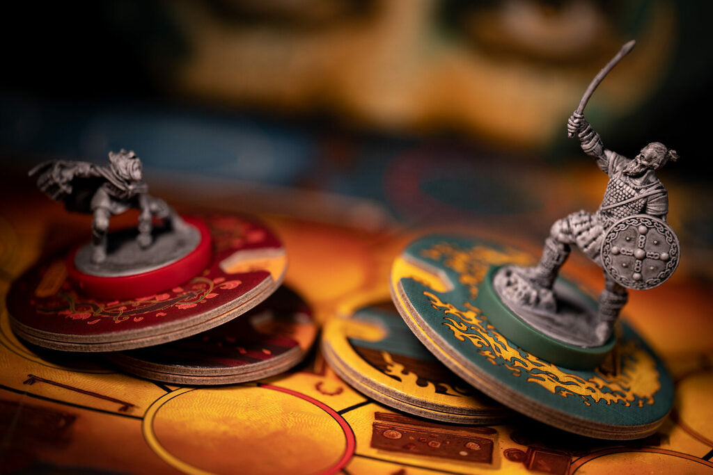 Two unpainted miniature fantasy warrior game figures standing on decorative round bases among blurred game tokens on a tabletop.