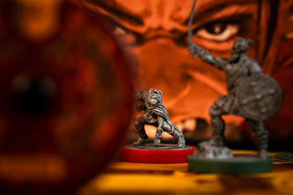 Two unpainted grey tabletop gaming miniatures on colored bases, with a blurred red shield in the foreground and a warm orange fantasy face backdrop.