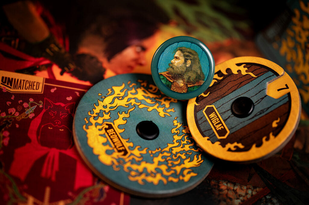 Three circular collectible tokens or badges featuring fantasy artwork, including a central round badge with a bearded character and two larger token discs with blue, yellow, and brown designs, arranged on a dark red illustrated surface.