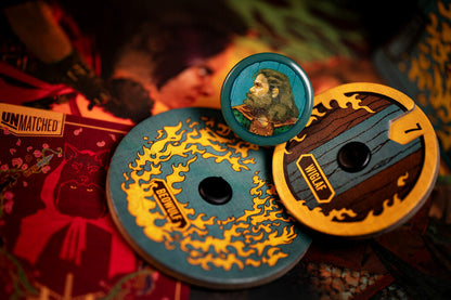 Three circular collectible tokens or badges featuring fantasy artwork, including a central round badge with a bearded character and two larger token discs with blue, yellow, and brown designs, arranged on a dark red illustrated surface.