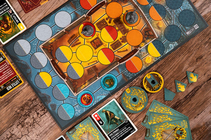 A top-down view of a board game setup on a wooden table, showing a blue and gray game board with circular path markers, a central play area with yellow and orange sections, cards, tokens, and dice.